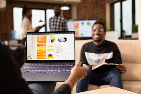 African american entrepreneur holding laptop showing company analytics, reviewing marketing strategy with male coworker. Selective focus on device held by businessperson, showing performance graphs.の写真素材