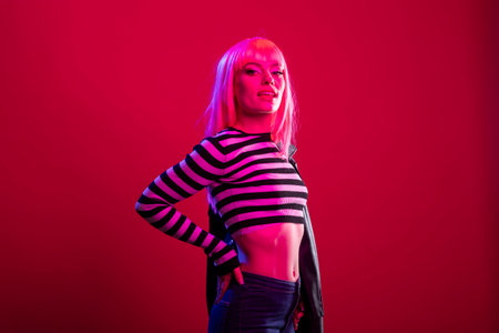 Female model with striped outfit and pink wig stands in calm yet assertive pose against isolated background, exuding vibrant energy. Glamorous lady with hand on waist, radiating confidence in studio.の写真素材