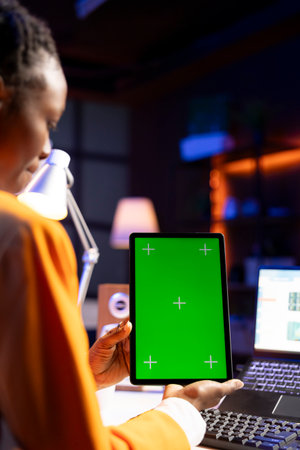 Woman sitting on chair at home office desk in neon illuminated apartment holding green screen tablet while working. Person looking at business graphs on laptop and at mockup deviceの写真素材