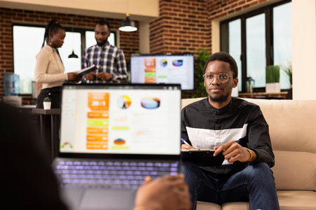 African american manager with laptop explains business plans and solutions to young male employee. Black businessman sitting on sofa with clipboard, attentively listens to coworker during meeting.の写真素材