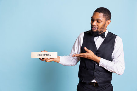 African american hotel porter presenting a wall sign for the reception area, showing an indicator to help guests in finding the front desk upon their arrival at holiday resort.の写真素材