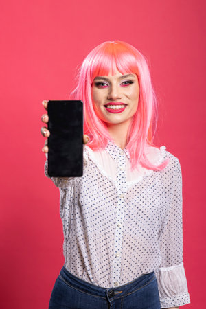 Caucasian woman with pink hair holding smartphone with blank screen, presenting copy space mockup display. Young female in casual wear showing mobile device against isolated background.の写真素材