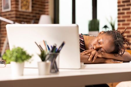 Overworked black businesswoman falls asleep at desk in home office after completing numerous tasks on laptop. Exhausted remote employee naps while seated, recovering from long hours of intense work.の写真素材
