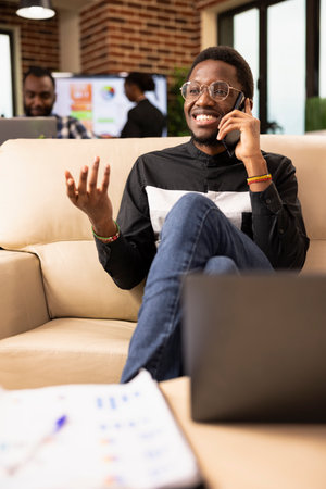 African businessman seated on sofa in brick wall office, enjoying a call with cellphone to his ear. Cheerful black male employee relaxing on the couch, speaking with friends on mobile device.の写真素材