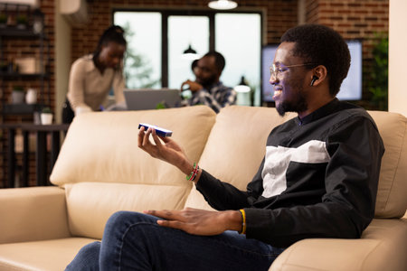 Young businessman seated on couch projects digital content in modern workspace. Behind him, black female manager and team collaborate, reflecting innovation and corporate teamwork.の写真素材