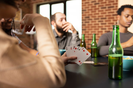 Closeup of woman playing cards with diverse friends, sharing beverages and snacks at home. Happy group playing poker at table with selective focus on deck of cards held by caucasian female individual.の写真素材