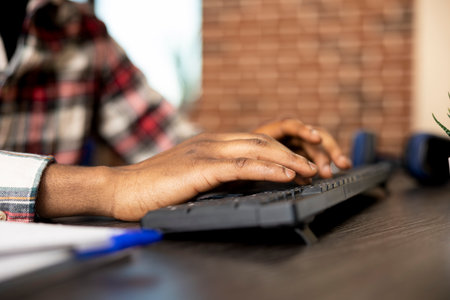 Closeup of african american individual using keyboard and updating company financial documents on desktop pc. Black man seated at home office desk, typing data in computer, preparing business project.の写真素材