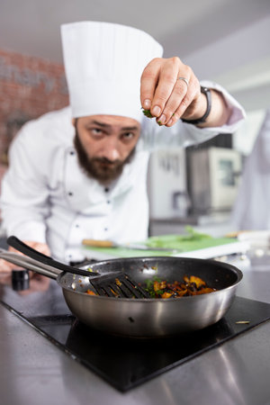 Professional chef in a modern kitchen carefully sprinkles fresh herbs onto a dish, enhancing the flavors with parsley, rosemary and basil. Modern workspace filled with fresh produce and spices.の写真素材