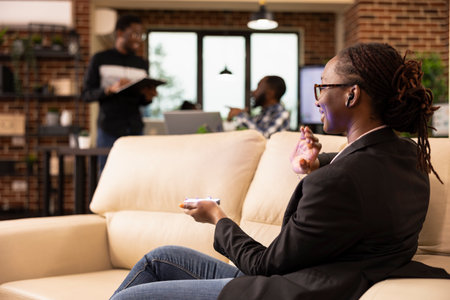 Black female entrepreneur using futuristic device to communicate with holographic interface and present data in tech forward workspace. African american team in background symbolizes startup synergy.の写真素材