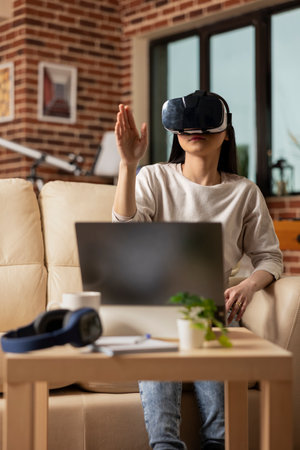 Remote female manager sitting in living room, using vr headset for immersive business planning. Young woman entrepreneur using futuristic tech, checking financial information and online presentations.の写真素材