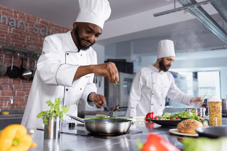 African american chef cooking delicious meals with rosemary or basil in a restaurant kitchen, sprinkles herbs leaves on the stove. Young cook using fresh produce and raw materials.の写真素材