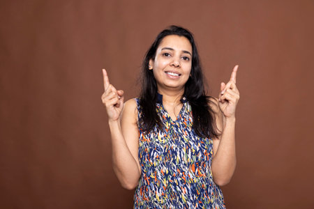 Joyful female model stands against brown background, smiling and pointing upward with both index fingers. Pretty indian woman drawing attention to space above her head for product or ad placement.の写真素材