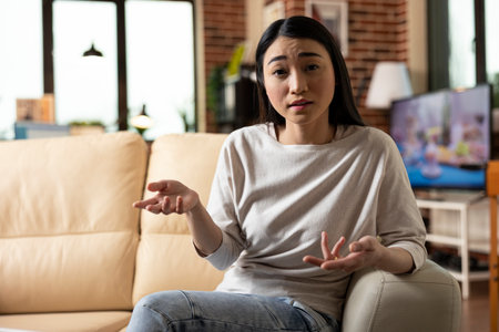 Focused asian female entrepreneur seated on couch, speaking toward camera while recording business presentation for potential investors. Modern remote work and startup concept.の写真素材