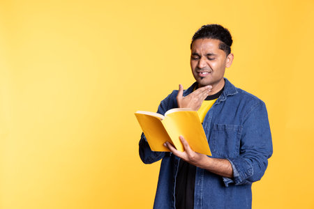 Joyous indian nerd enthusiast reading book for pupils during class, standing against orange background. Happy pleased man holding novel, telling story and relaxing with the lecture in studio.の写真素材