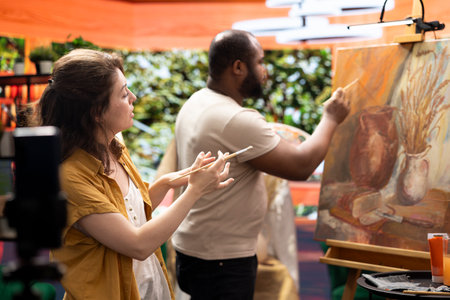 Woman painter filming her colleague during his creative process and explaining the painting technique for online tutorial. Diverse artists creating virtual classroom to teach art. Digital education.の写真素材