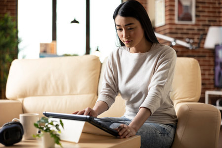Asian businesswoman lifts laptop lid to begin remote work day in brick wall apartment. Female freelancer with digital device on table, preparing for research, planning and skill building.の写真素材