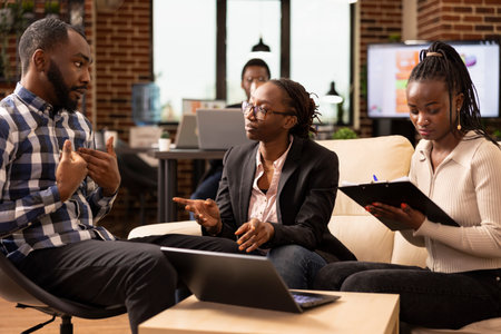 African american professionals discuss marketing strategies and business solutions, with female manager wearing eyeglasses providing updates to coworkers in cozy office lounge.の写真素材