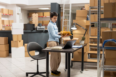 Woman depot worker preparing boxes for shipment in local brand supply room, managing the orders on a laptop. Fulfillment center focused on retail operations and e commerce distribution.の写真素材