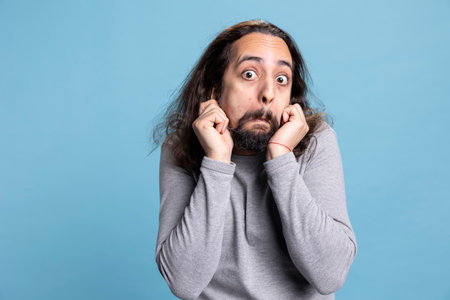 Joyful bearded man with long curly hair strikes a silly pose showing a carefree attitude. Playful behavior and comical face highlight his humorous personality in this studio against blue background.の写真素材