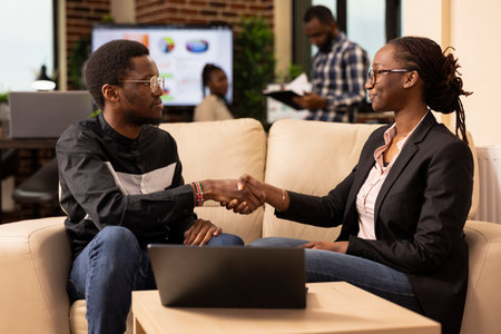 African American male and female coworkers shake hands at office couch, with laptop on table. Professional black man and woman sealing successful business project deal in modern workspace.の写真素材