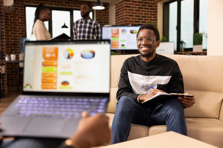 Two business professionals meet in office to share ideas and collaborate on project task. African american sales manager with laptop presents commercial offer to startup company client seated on sofa.の写真素材