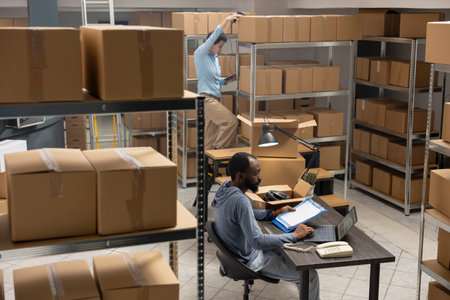 Business owner overseeing product handling and shipping systems inside a compact logistics center, retail activity. Black warehouse worker prepares boxes for shipment from supply room.の写真素材