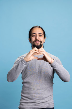 Romantic person showing a heart shape sign against blue background, doing a symbol of affection and passion in studio. Young adult acting playful in the art of seduction on camera.の写真素材