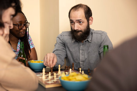 Selective focus on caucasian man holding chess piece, concentrating on his female opponent during their game. Bearded white guy sits at table with snacks and chessboard, planning his next move.の写真素材
