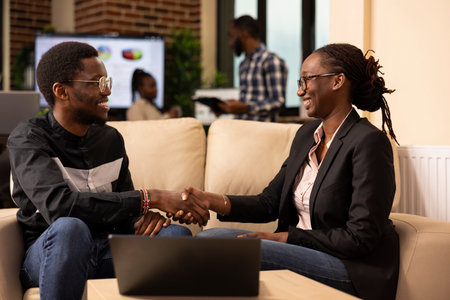 Happy businessman and businesswoman shaking hands near laptop while analyzing performance metrics. African american colleagues seated on couch, showcasing teamwork and excitement in marketing agency.の写真素材