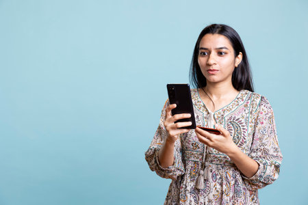 Upbeat person adding banking credentials on her mobile phone, confirming payment and money transfer on a website. Young indian adult buying online using card against studio background.の写真素材