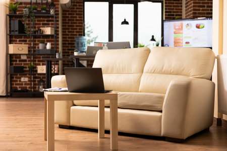 Vacant brick wall workspace neatly organized for employees at startup company. Empty cozy sofa with laptop nearby on table, highlighting comfort and productivity in modern office.の写真素材