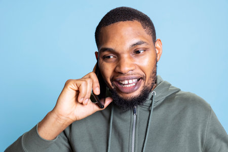 African american young adult enjoys talking to his friends on phone call using smartphone, poses against studio background. Relaxed guy catching up with mates during telephone call.の写真素材