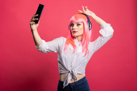 Caucasian woman wearing wireless headphones and taking playful selfies against vibrant pink backdrop for her social media. Stylish young lady dressed casually and snapping shots with her cellphone.の写真素材