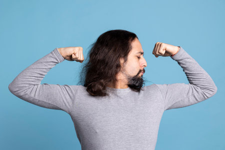 Athletic young guy flexing his arms muscles against blue background, showing off his toned fit physique in front of the camera. Fitness motivation and determination from a strong man.の写真素材