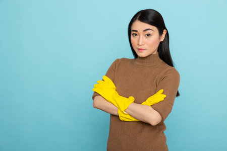 Portrait of female janitor wearing yellow gloves, standing confidently with crossed arms, ready to provide cleaning services. Asian housekeeper posing against blue studio background.の写真素材