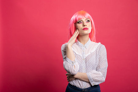 Young female individual with hand on cheek, looking upward in thoughtful contemplation. Pensive woman with pink hair stands against isolated background, appearing focused and reflective.の写真素材