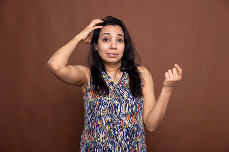 Puzzled adult woman scratching her head and looking at camera with troubled expression in brown background studio. Pretty indian woman appearing confused and indecisive, unsure about something.の写真素材