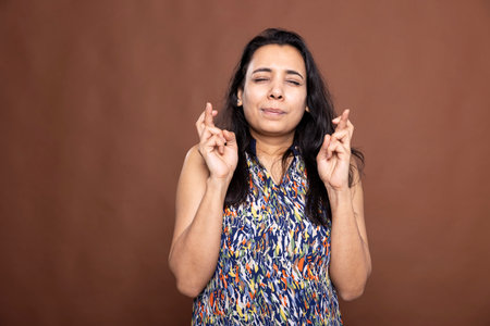 Indian woman stands with eyes closed and fingers crossed, quietly hoping for favorable result. Female individual with pleading expression, silently wishing for good news or miracle.の写真素材