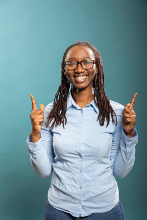 Smiling african american woman points upward with both index fingers, highlighting space for promotional content. Black female model in blue shirt, looking at camera and gesturing above.の写真素材