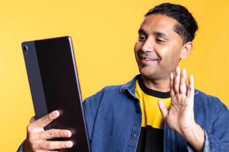 Indian person waving at the online video call on tablet against orange background, saluting his friends on web conference meeting in studio. Young guy in casual attire being friendly.の写真素材