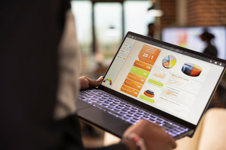 African american manager standing with laptop in hand, reviewing company analytics and charts in...