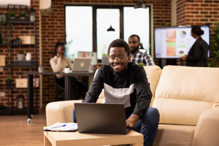 Black male marketing expert works from cozy lounge area, reviewing business insights. African American man sits on comfortable sofa with laptop and clipboard, analyzing startup data in modern office.の写真素材