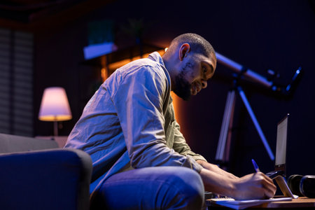 Black male freelancer taking notes on his textbook in order to solve his tasks, doing research and...