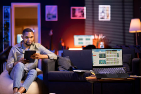 Young Black man relaxes on sofa and uses PC to track forex trades, analyzing stock market graphs for...