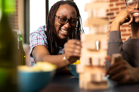 Happy black woman filled with laughter, pulling block during wooden tower game, as male friend watches nearby with a smile. Game day excitement builds during their cozy home gathering.の写真素材