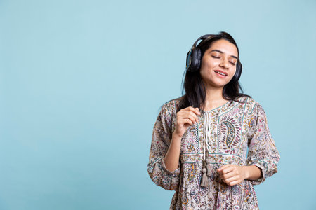 Indian woman enjoying groovy music on headphones and dancing, having fun moving on rhythm against blue background. Pleased young adult does groovy movements wearing audio headset.の写真素材