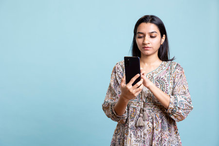 Indian woman happily scrolling on phone to check social media feed, amazed by something. Young relaxed person browsing the internet websites, texting friends against studio background.の写真素材