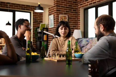 Smiling young woman sits with arms crossed during chess match, calmly observing her male friends play. Caucasian lady in casual pose, gazing at camera amid board game session in brick wall room.の写真素材