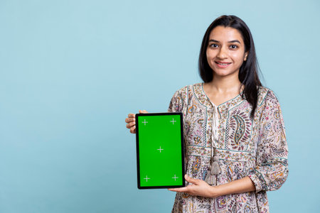 Portrait of indian woman doing marketing ad with green screen device, standing against blue background. Smiling young adult feeling positive on an advertisement in front of the camera.の写真素材