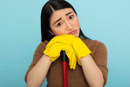 Frustrated asian woman in yellow rubber gloves leans on mop, showing displeasure and signs of...